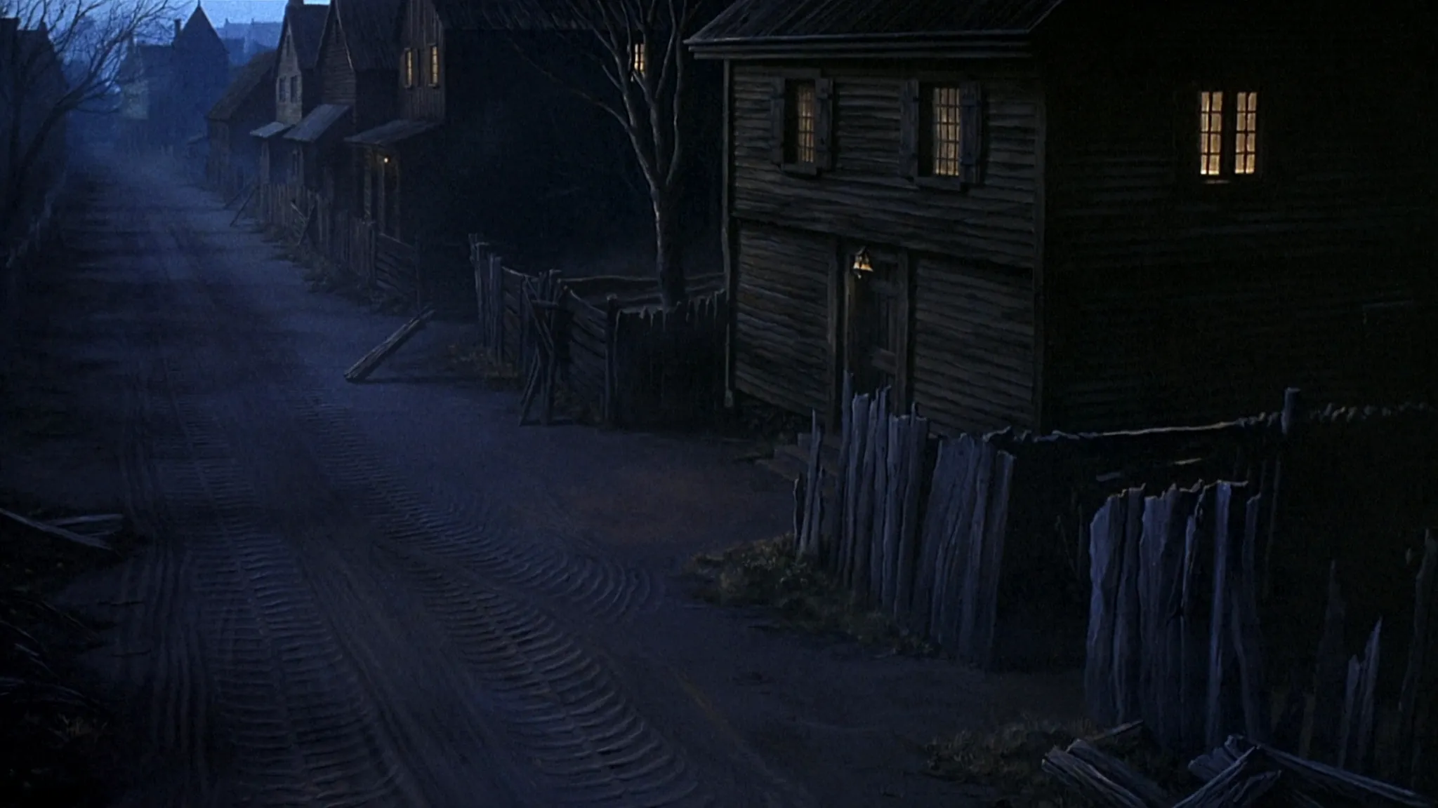 A quiet Salem village lane at dusk with lantern light and long shadows.